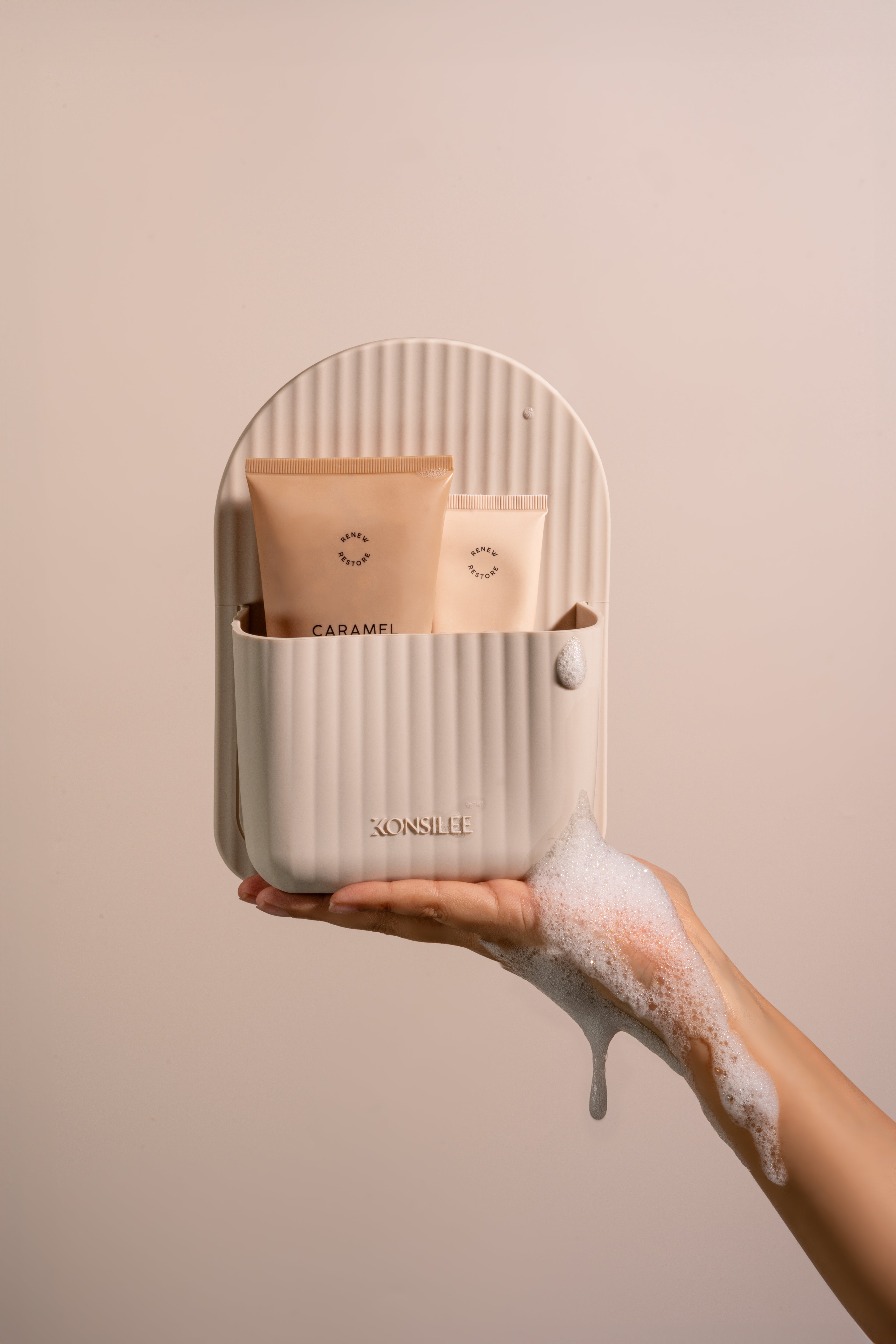 A soft-brown, fluted silicone shower caddy, held in a person’s soapy hand. The organiser has an arch shape and an embossed Konsilee logo. It contains two tan skincare tubes. The background is a solid peach colour.