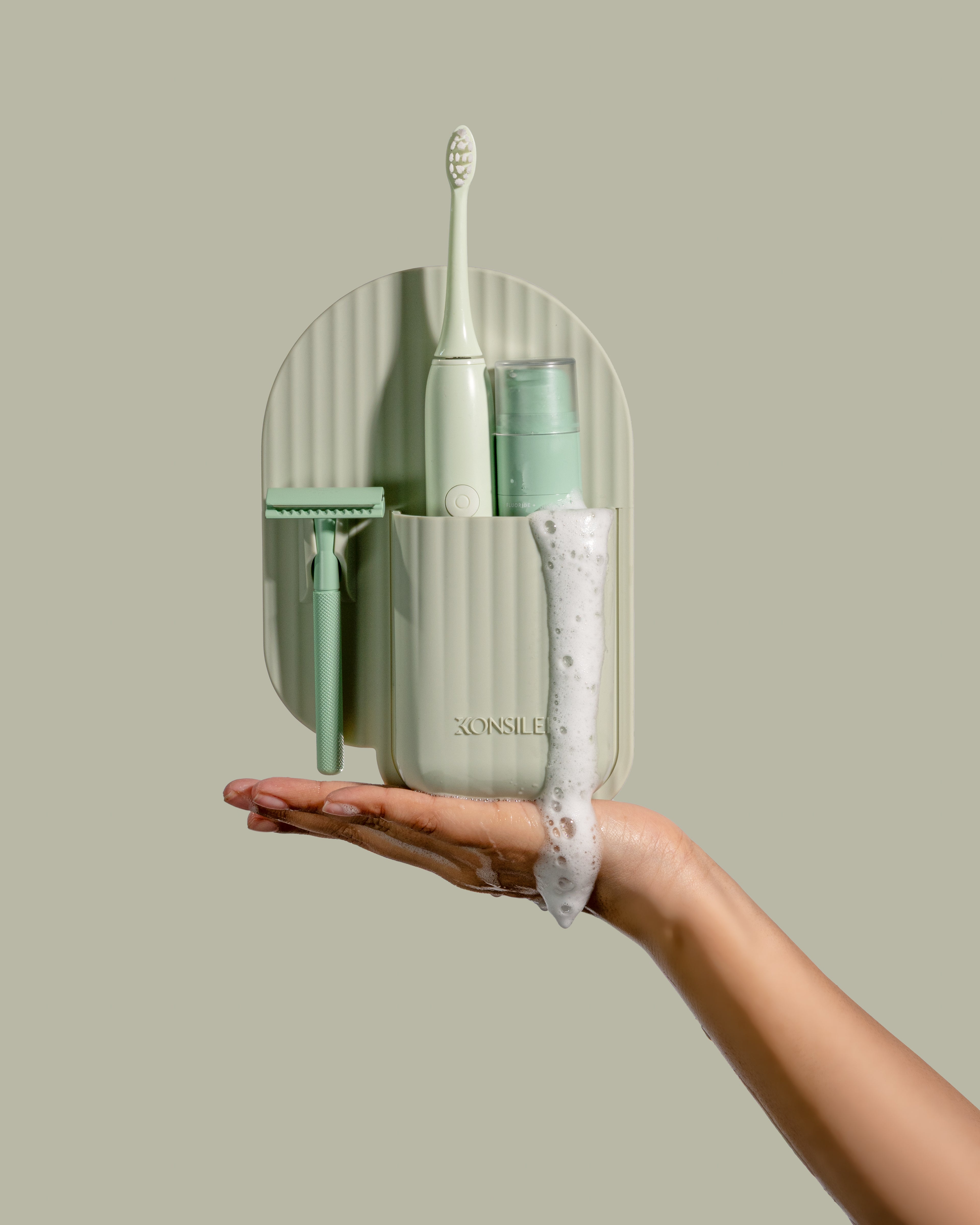 A soft-green, fluted silicone toothbrush and razor caddy, held in a person’s soapy hand. The organiser has an arch shape and an embossed Konsilee logo. It contains a green razor, electric toothbrush and toothpaste bottle. The background is a solid light-green shade.