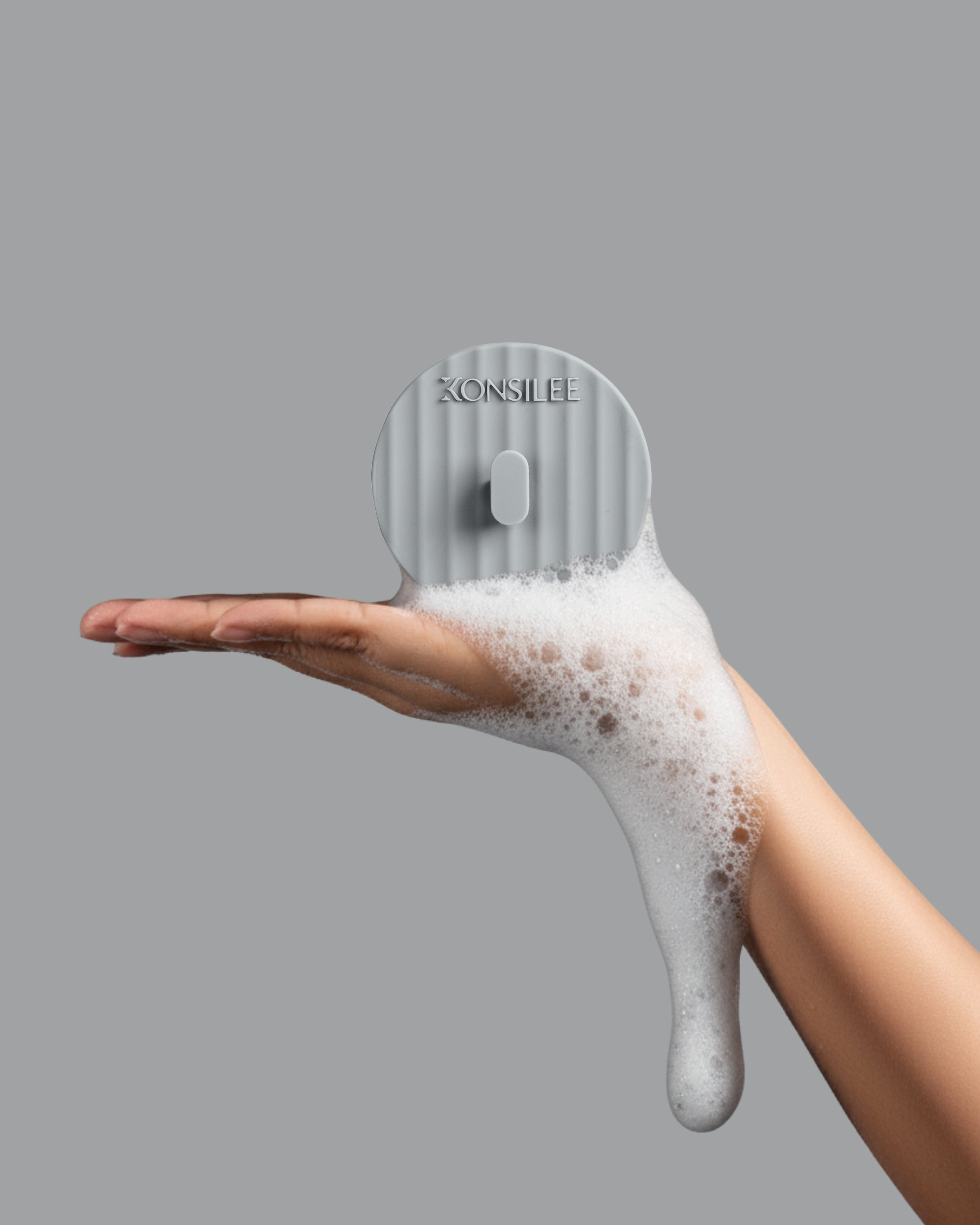 A light-grey, fluted silicone shower hook, held in a person’s soapy hand. The hook is a circle shape and has an embossed Konsilee logo. The background is a solid light-grey shade.