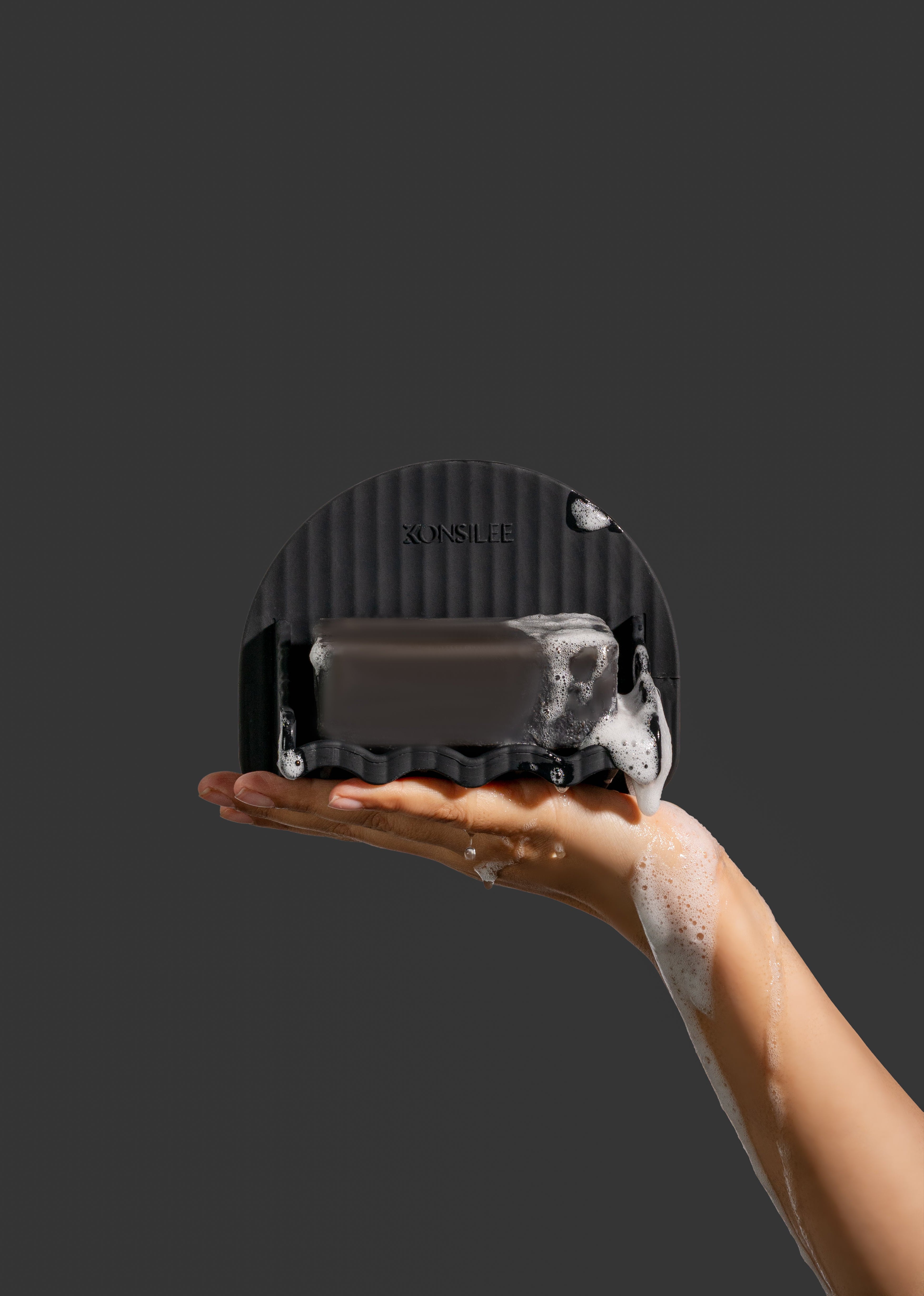 A black, fluted silicone soap dish with an arched silhouette and a wavy drainage ledge. It features an embossed Konsilee logo and is held in a soapy hand against a solid dark charcoal background.